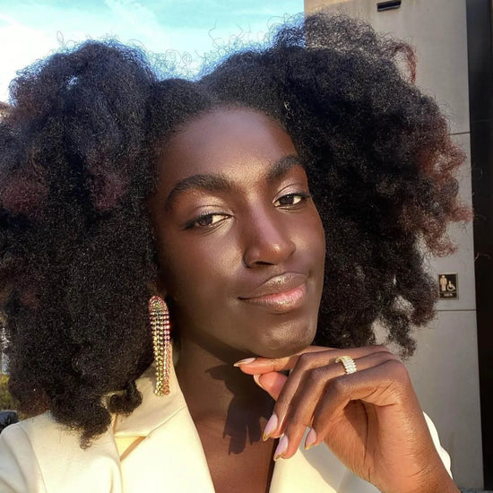 A person with curly hair poses outdoors, wearing colorful earrings and a ring.